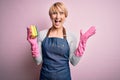 Young blonde cleaner woman with short hair wearing apron and gloves holding scrub scourer very happy and excited, winner