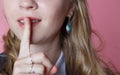 young blonde curly haired girl holds her finger to her lips on a pink background close-up, concept of secret and silence. Royalty Free Stock Photo