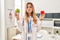 Young blonde doctor woman holding heart and green apple puffing cheeks with funny face Royalty Free Stock Photo