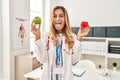 Young blonde doctor woman holding heart and green apple sticking tongue out happy with funny expression Royalty Free Stock Photo