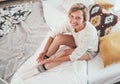 Young blonde female dressed in pajama sitting in the cozy bed with pillows and sincerely smiling and gazing at the camera. Staying