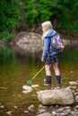 A young blonde girl with backpack and fishing net wades in the shallow water of a river