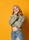 Young blonde girl in torn jeans with finger on her cheek smiles and thinks over or dream about something on yellow background