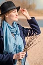 Young blonde girl in dress, coat and hat in spring on beach, under rays of spring sun with willow branches, smiling Royalty Free Stock Photo