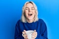 Young blonde girl drinking a cup of coffee smiling and laughing hard out loud because funny crazy joke Royalty Free Stock Photo