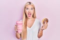 Young blonde girl eating doughnut and drinking coffee angry and mad screaming frustrated and furious, shouting with anger looking Royalty Free Stock Photo