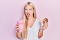 Young blonde girl eating doughnut and drinking coffee angry and mad screaming frustrated and furious, shouting with anger Royalty Free Stock Photo