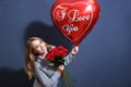 Young blonde girl in a gray sweater holding a bouquet of red roses and a heart balloon. Happy valentine day concept. Royalty Free Stock Photo