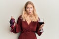 Young blonde girl holding branch of fresh grapes and red wine afraid and shocked with surprise and amazed expression, fear and Royalty Free Stock Photo