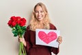 Young blonde girl holding heart draw and bouquet of roses sticking tongue out happy with funny expression