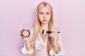 Young blonde girl holding makeup bronzer and brush puffing cheeks with funny face