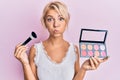 Young blonde girl holding makeup brush and blush puffing cheeks with funny face