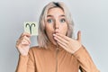 Young blonde girl holding paper with aries zodiac sign covering mouth with hand, shocked and afraid for mistake