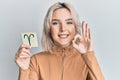 Young blonde girl holding paper with aries zodiac sign doing ok sign with fingers, smiling friendly gesturing excellent symbol