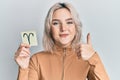 Young blonde girl holding paper with aries zodiac sign smiling happy and positive, thumb up doing excellent and approval sign