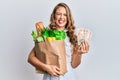 Young blonde girl holding paper bag with groceries and pounds sticking tongue out happy with funny expression