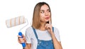Young blonde girl holding roller painter serious face thinking about question with hand on chin, thoughtful about confusing idea Royalty Free Stock Photo