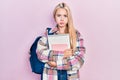 Young blonde girl holding student backpack and books puffing cheeks with funny face