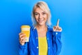 Young blonde girl holding take away coffee smiling with an idea or question pointing finger with happy face, number one