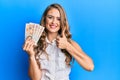 Young blonde girl holding united kingdom pounds smiling happy and positive, thumb up doing excellent and approval sign