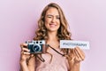 Young blonde girl holding vintage camera and paper with photography word paper winking looking at the camera with sexy expression,