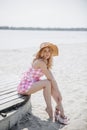 Young blonde girl look like a Barbie doll in pink mini dress and wide brim hat sitting barefoot on beach Royalty Free Stock Photo