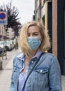 A young blonde girl with a purposeful look in a medical protective mask on a European street. In a denim jacket