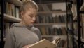 Young blonde girl reading a book standing between bookshelves in the library