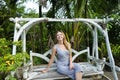 Young blonde girl riding on swing in exotic garden, palms in background.