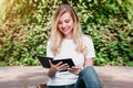 student girl sits on a bench, reads a book and smiles in a park on a background of trees and bushes Royalty Free Stock Photo