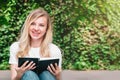 Young student girl sits on a bench, reads a book and smiles in a park on a background of trees and bushes Royalty Free Stock Photo