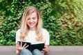 Young blonde student girl sits on a bench, reads a book and smiles in a park Royalty Free Stock Photo