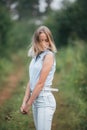 A young blonde girl stands on a forest road with flowers in her hands and turns her head.