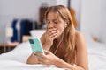 Young blonde girl using smartphone and earphones tired lying on bed at bedroom