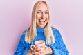 Young blonde girl wearing winter sweater and drinking a cup of hot coffee smiling and laughing hard out loud because funny crazy Royalty Free Stock Photo