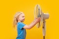 Young blonde girl with windblown hair near fan on yellow background. Fair-haired child near blower in hot weather Royalty Free Stock Photo