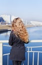 Young blonde model girl with long curly hair standing on the bridge and looking at the river and city in windy day