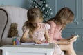 Young blonde mom sitting on couch in pink blouse writing plans. Christmas tree. Daughter raws pictures in the album with