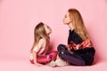 Young blonde mother and her daughter in glossy leggings and sunglasses sit face to face on floor going to sending kiss Royalty Free Stock Photo