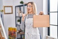 Young blonde painter woman holding wooden case at art studio pointing thumb up to the side smiling happy with open mouth Royalty Free Stock Photo