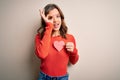 Young blonde romantic girl holding red heart paper shape over isolated background with happy face smiling doing ok sign with hand Royalty Free Stock Photo