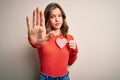 Young blonde romantic girl holding red heart paper shape over isolated background with open hand doing stop sign with serious and Royalty Free Stock Photo