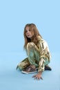 A  young blonde serious girl wearing in a golden jumpsuit sits on blue background looks aside and thinks about something. The Royalty Free Stock Photo