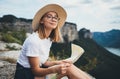 Young blonde tourist holds in hands map of landscape and looks views top of mountain landscape, cute smiling girl in hat