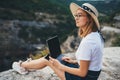 Young blonde tourist looks at laptop screen and enjoys mountain landscape, ÃÂute girl in hat and glasses works on wireless