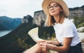 Young blonde tourist planning trip on map and sitting on top of mountain landscape, cute girl in hat and glasses is relax