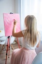 Young blonde woman artist with palette and brush sitting and painting abstract pink picture on canvas near window at