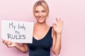 Young blonde woman asking for women freedom holding paper with my body my rules message doing ok sign with fingers, smiling