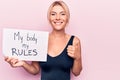 Young blonde woman asking for women freedom holding paper with my body my rules message smiling happy and positive, thumb up doing