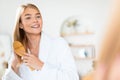 Young blonde woman in bathrobe enjoys grooming with hairbrush indoor Royalty Free Stock Photo